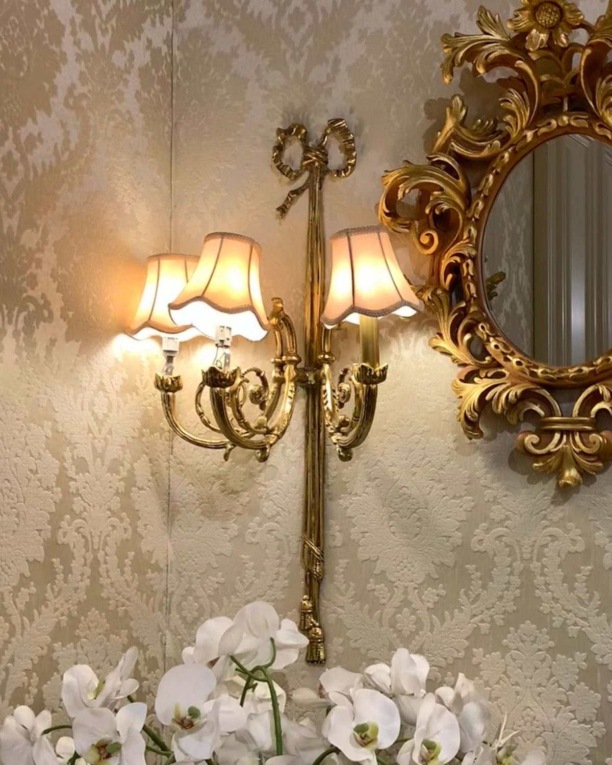 Cream damask wallpaper with a gilt mirror, brass sconce and white orchids