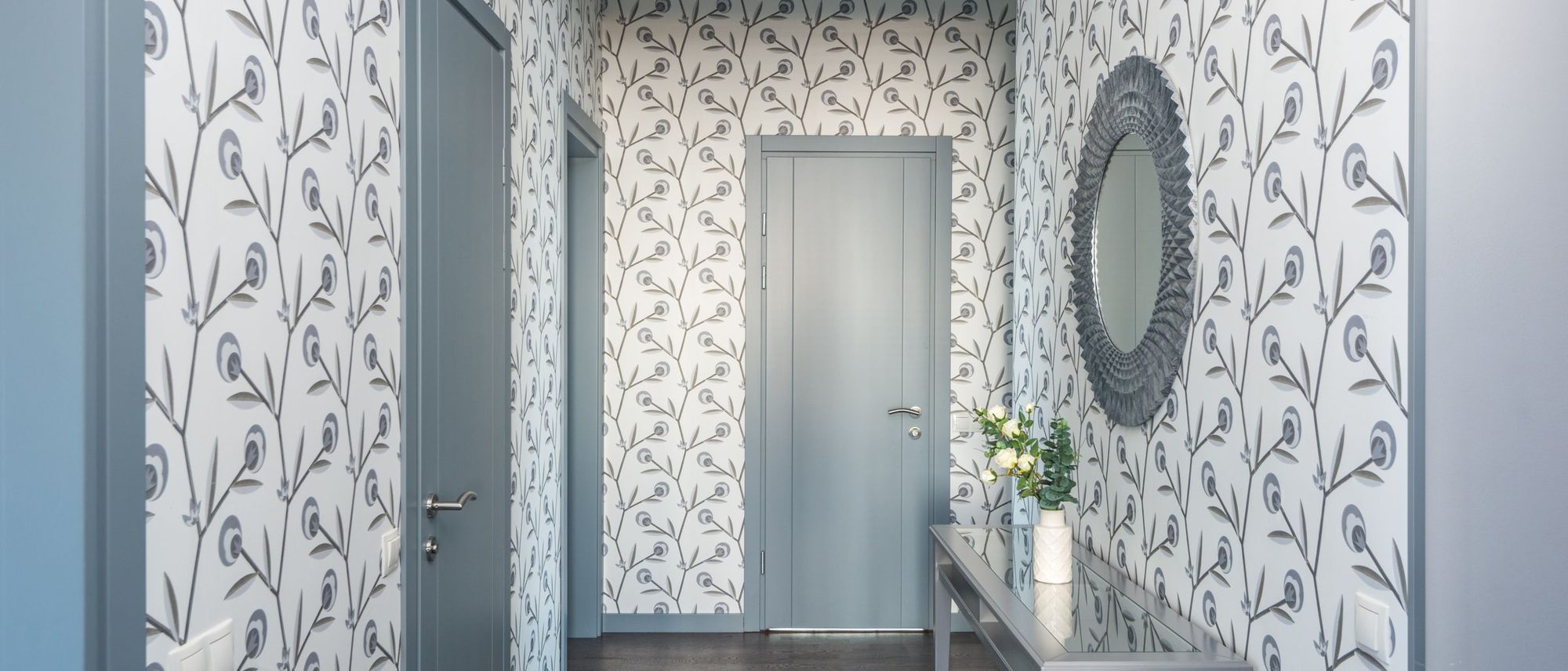 Long residential hallway with delicate white botanical wallpaper, dark wood floor and a round mirror